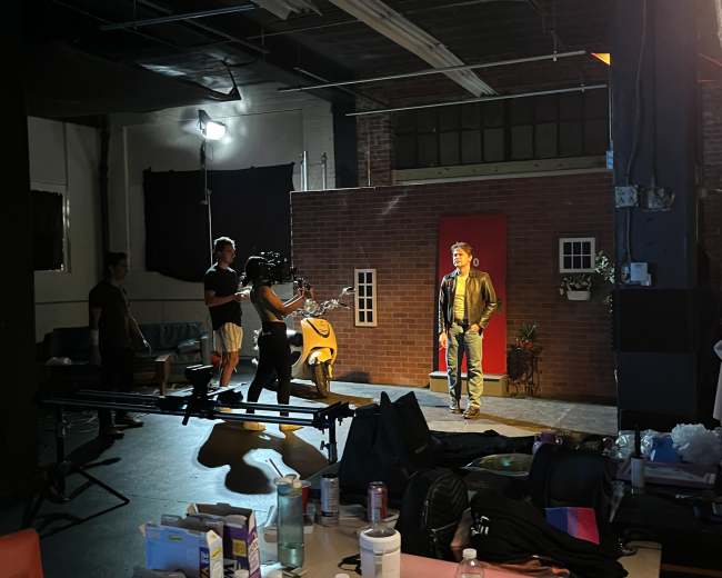 A film crew sets up in a studio, with a lead actor standing in front of a brick wall backdrop while a camera is positioned nearby.