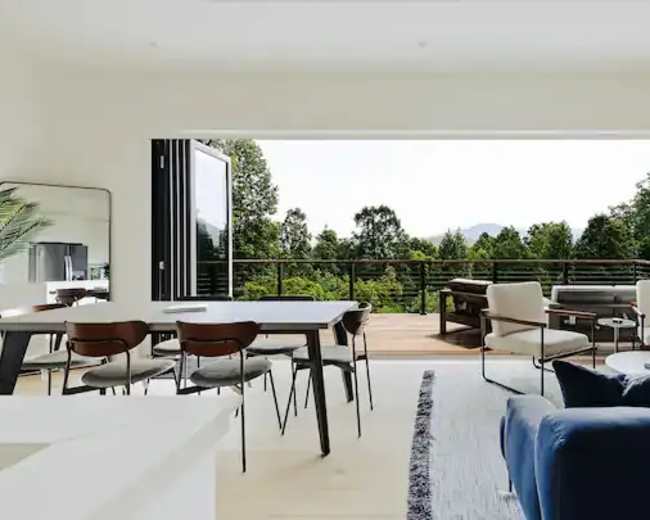 A modern living space with a dining area, a seating area, and large sliding glass doors that open to a deck surrounded by trees.