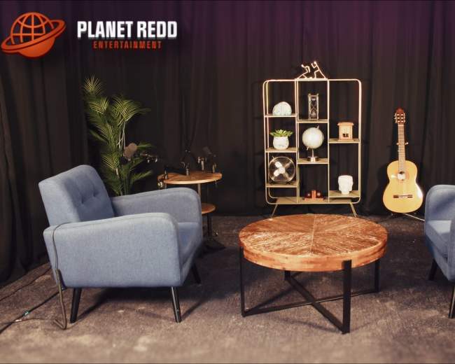 A cozy set features two blue chairs facing a wooden coffee table, flanked by plants and a guitar on a shelf in the background.