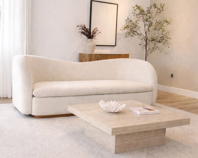 The image shows a light-colored, modern living room featuring a curved sofa, a simple wooden coffee table, a decorative bowl on the table, and greenery in a corner.