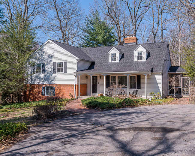 A two-story white house with a black roof and a brick front porch, surrounded by trees and a gravel driveway.