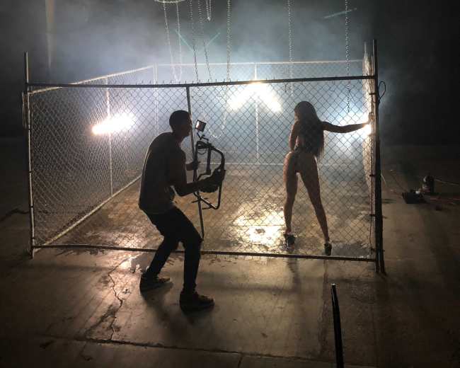 A person holds a camera while filming a woman in a fenced-off area with dramatic lighting and fog.