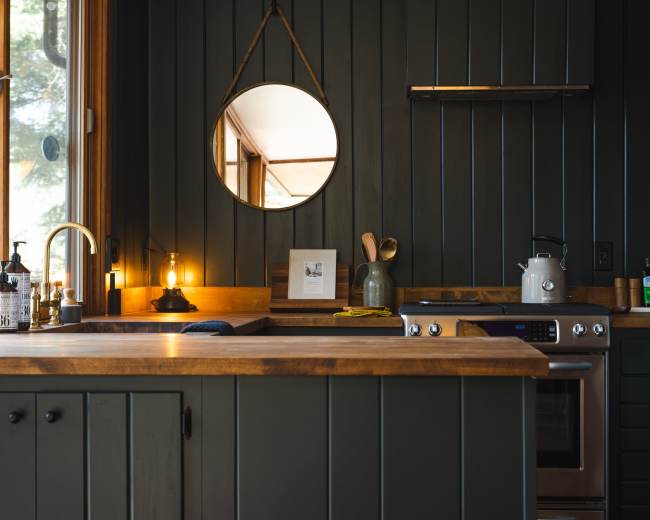 The image shows a modern kitchen with dark green paneling, a wooden countertop, and a round mirror mounted on the wall.