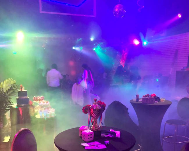 A dimly lit party scene features tables with decorated snacks and a foggy atmosphere enhanced by colorful lighting and silhouettes of people mingling.