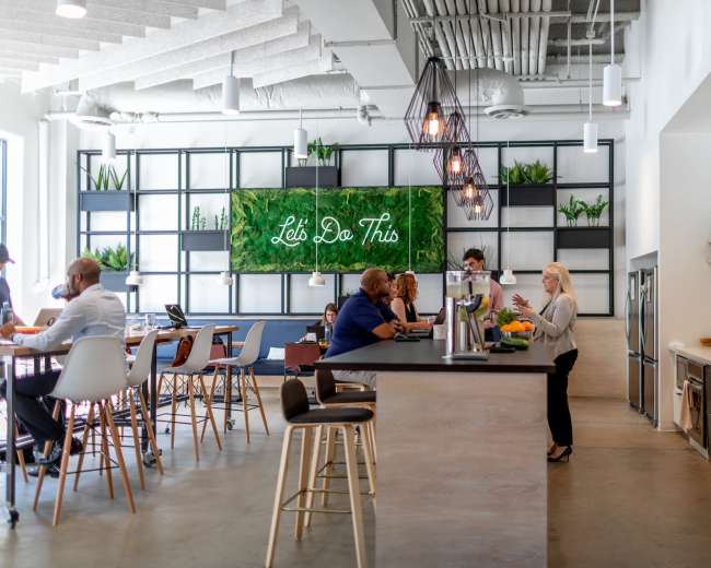 A modern office space with a kitchenette, a bar-style counter, and seating areas where people are engaged in conversation and work activities.