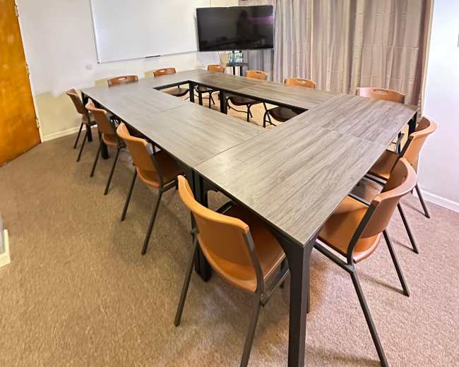 The image shows a rectangular conference room with a large wooden table surrounded by orange chairs and a TV monitor on the wall.