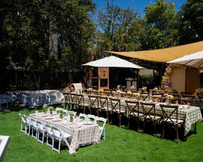 A large outdoor dining setup features multiple tables with checkered tablecloths, surrounded by shaded areas created by umbrellas and a fabric canopy.