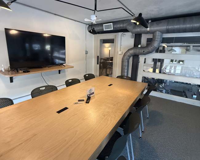 The image shows a modern conference room featuring a large wooden table surrounded by black chairs, with a wall-mounted television and exposed ducts on the ceiling.