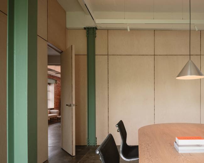 A modern meeting room features a round wooden table, sleek chairs, and a pendant light, with green beams and wood-paneled walls.