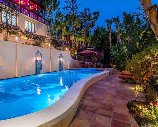 A curved swimming pool with three wall fountains is surrounded by lush greenery and outdoor seating, illuminated by soft lights at dusk.