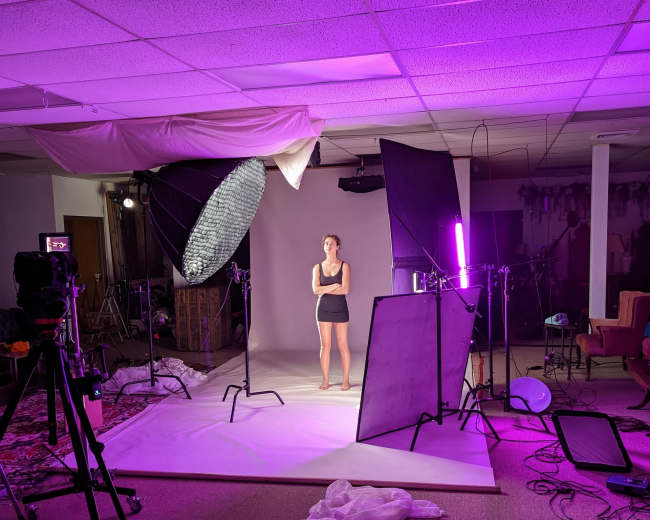 A model stands on a light backdrop in a studio illuminated by purple and softbox lighting, surrounded by various photographic equipment.
