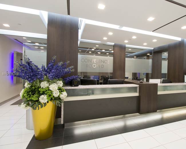 The image shows a modern reception area featuring a conference room sign, a sleek front desk, and large floral arrangements in yellow pots on either side.