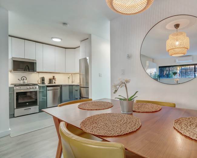 A dining table with woven placemats and a potted orchid is positioned in front of a kitchen with modern appliances and cabinetry.