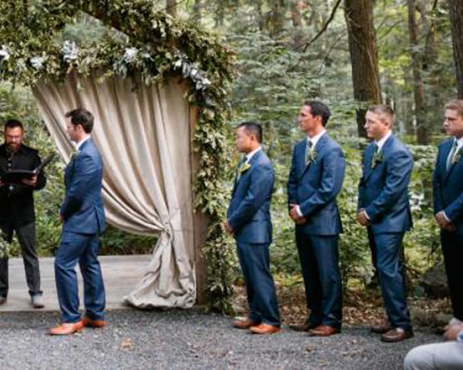 A wedding ceremony is taking place in a forested area under a floral arch, with the couple facing an officiant surrounded by their wedding party.