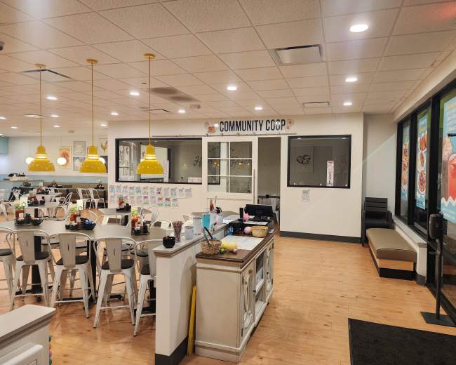 The image shows an interior of a communal dining area with a service counter, seating arrangements, and bright yellow pendant lights.