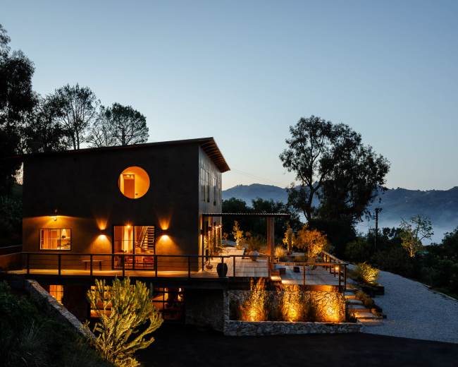 A modern two-story house with exterior lighting sits on a hillside, surrounded by trees and misty mountains at dusk.