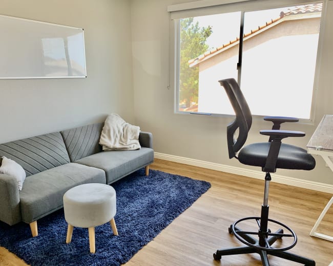 A minimalist office space featuring a gray sofa with a light blanket, a round stool, a swivel chair, and a desk by a window.
