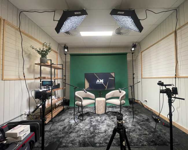 The image shows a podcast studio setup with two white chairs, a green backdrop, and multiple cameras and lighting equipment.