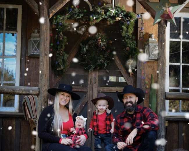 A family wearing matching red and black outfits poses on the steps of a cabin, with snow falling around them.