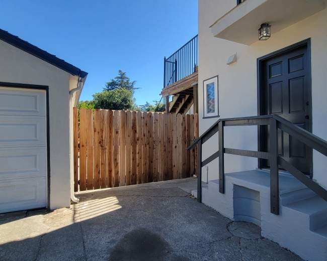 The image shows a pathway leading to a door next to a white garage and a wooden fence, with a staircase visible above.