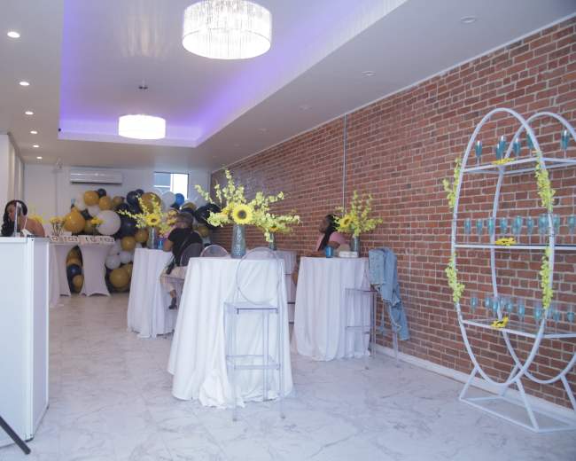 A smoothly arranged event space featuring white tables with decorations, a brick wall, and a balloon display in the background.