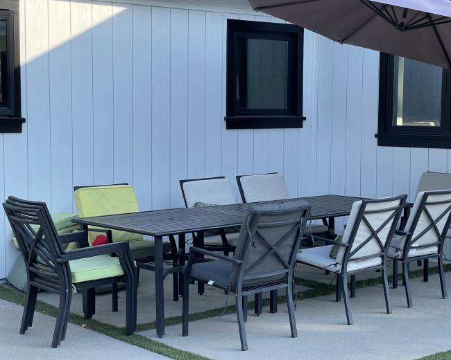 A rectangular outdoor dining table is surrounded by several chairs on a concrete patio, with an umbrella positioned nearby.