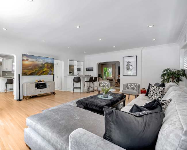 A modern living room features a sectional sofa, a coffee table, and a large television, with an open kitchen visible in the background.