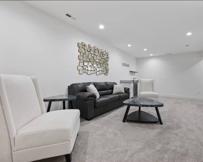 The image shows a modern living room with a dark sofa, two white armchairs, and a coffee table, all set against a light-colored wall with a decorative wall piece.
