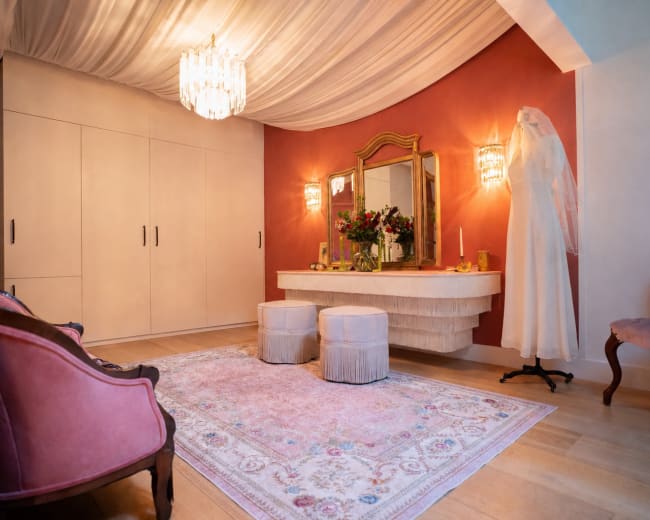 The image shows a well-decorated dressing area with a vintage vanity, two upholstered stools, a wedding dress on a mannequin, and warm lighting under a draped ceiling.