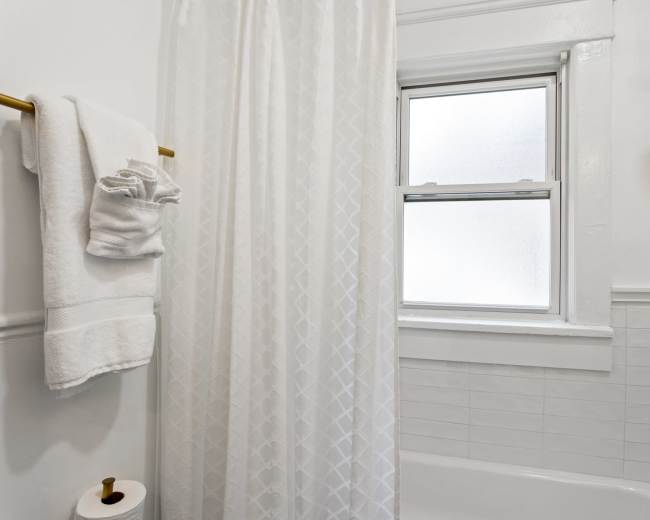 A bathroom features a white shower curtain, hanging towels, and a window above a bathtub.