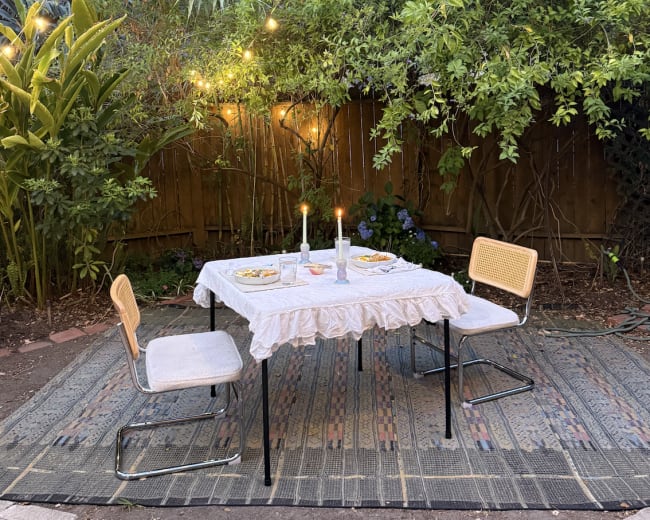 A table set for dinner with two chairs, candles, and plates, surrounded by greenery and fairy lights in an outdoor space.