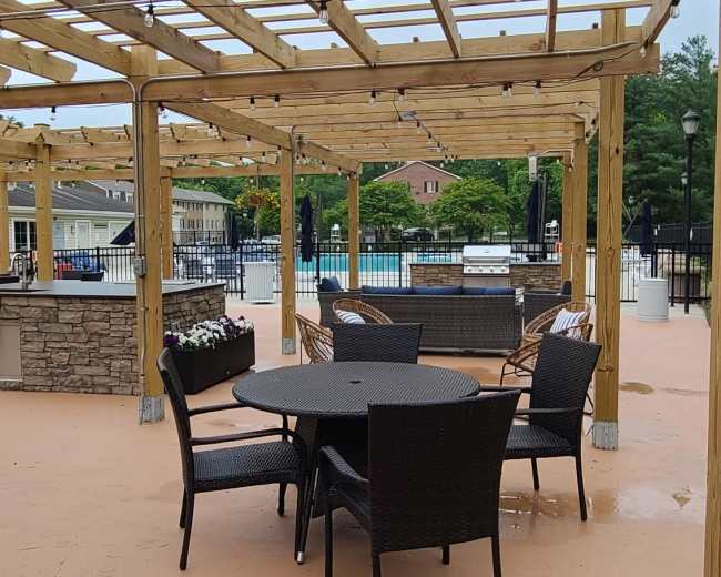 The scene features a wood-framed pergola with a seating area composed of a blue upholstered sofa and matching ottomans, surrounded by various outdoor chairs on a concrete surface.