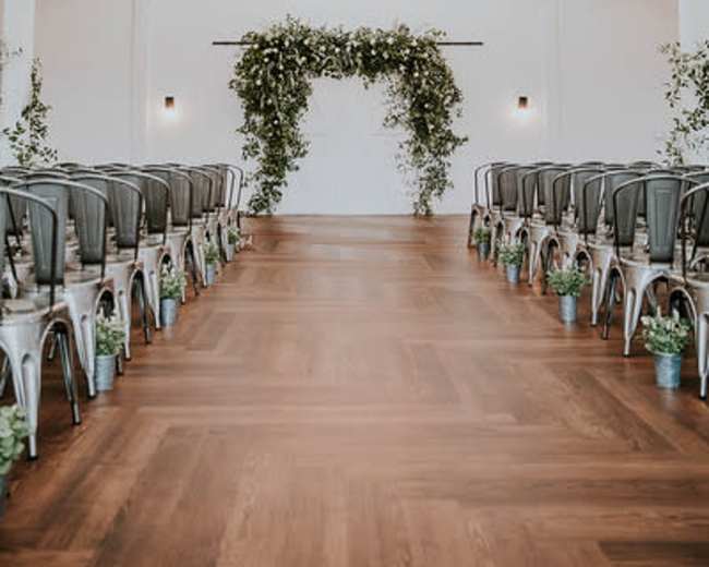 A spacious indoor venue is set up for a wedding ceremony, featuring rows of metal chairs facing a floral arch under two large, circular light fixtures.