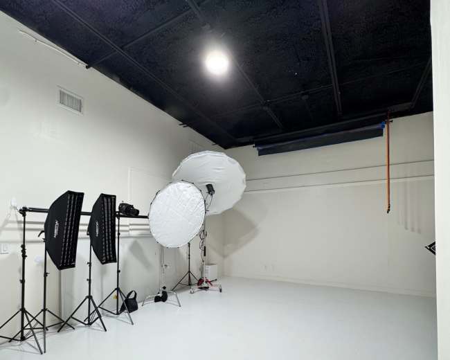 The image shows a spacious photography studio with white walls, a polished floor, and lighting equipment set up in one corner.