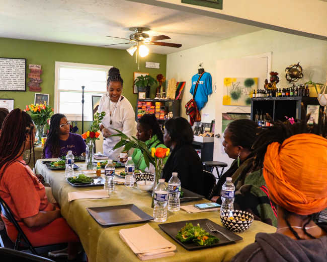 A group of eight people sit around a table with plates of food and water bottles while a woman in a white coat stands to address them in a cozy room filled with plants and art.