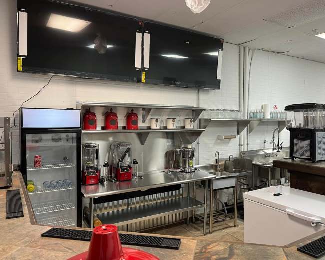A modern commercial kitchen area features stainless steel appliances, including blenders, a refrigerator, and a drink station, with two large flat-screen TVs mounted on the wall.