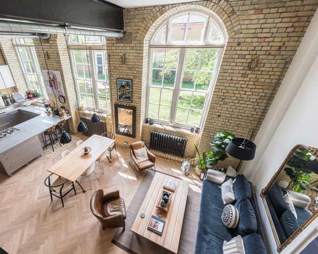 The image shows a spacious, modern living area with large windows, featuring a kitchen, dining table, and comfortable seating.