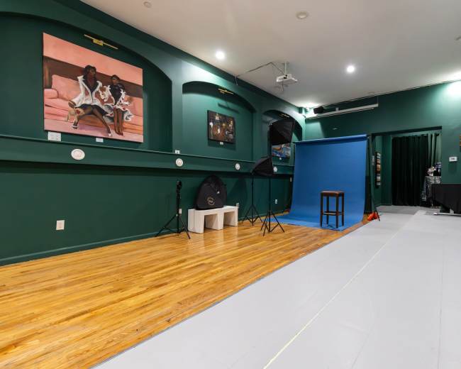 The image depicts a studio space with green walls, a wooden floor, a blue backdrop, and various lighting equipment arranged for photography.