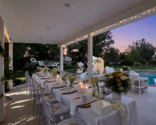 An elegantly set outdoor dining area features a long table adorned with flowers and decorations, overlooking a swimming pool and a colorful sunset in the background.