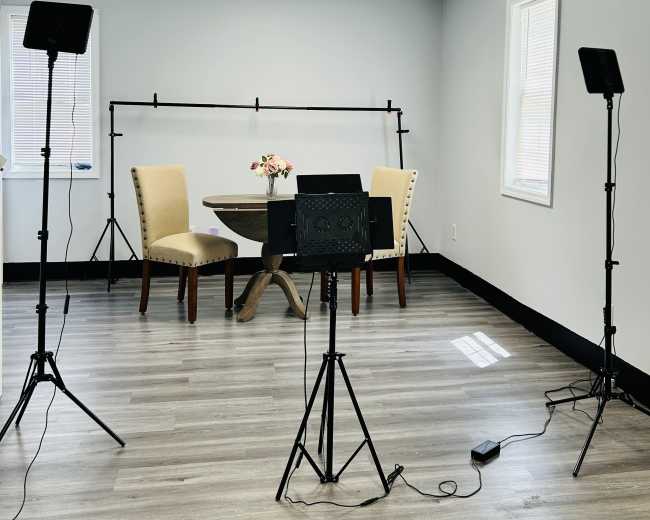 The image shows a well-lit interior room with a small round table and two chairs, flanked by lighting equipment and a flower arrangement on the table.