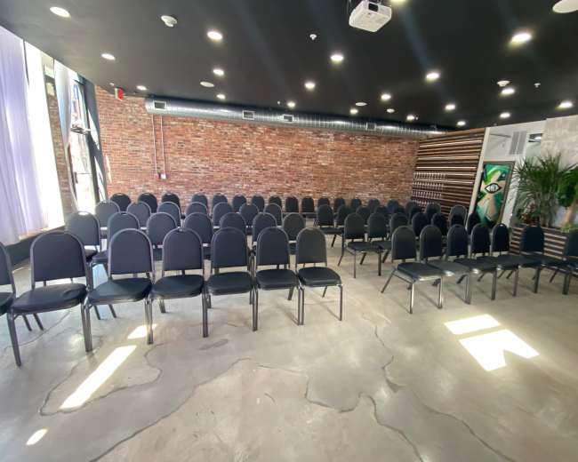 The image shows a room arranged with rows of black chairs facing a brick wall and large windows, with polished concrete flooring.