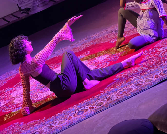 Two performers are engaged in a visual narrative on a red rug, with one sitting and reaching out while the other reclines, and a third person is speaking into a microphone in the background.