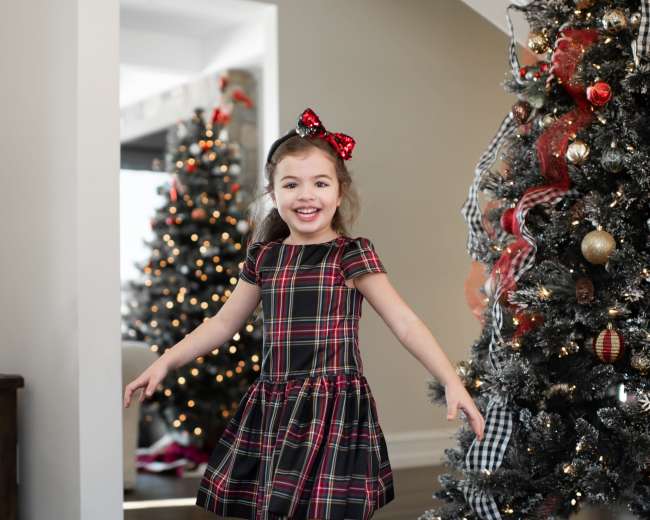 A young girl in a plaid dress and a red bow twirls in a room decorated for the holidays, with a Christmas tree in the background.