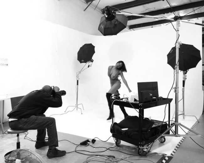 A photographer captures a model posing in a studio with white backdrops and lighting equipment.