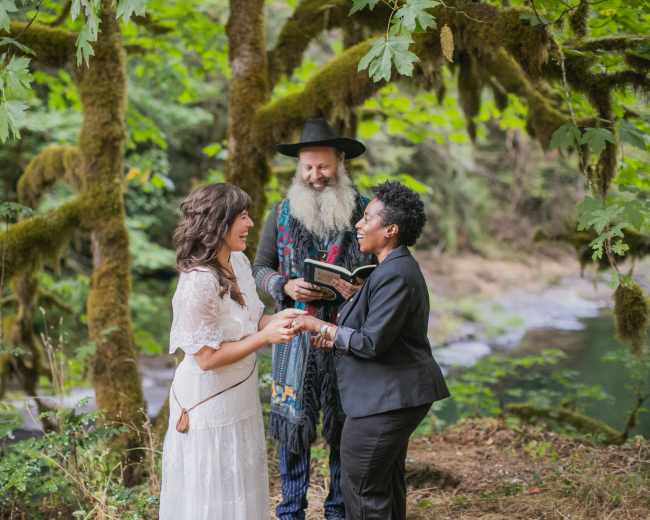 A couple stands hand in hand while a bearded officiant reads from a book in a forested area near a creek.