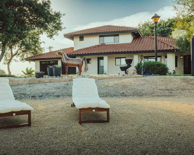 The image features two white lounge chairs on a gravel area in front of a modern, two-story house with a red tile roof and a horse sculpture in the yard.
