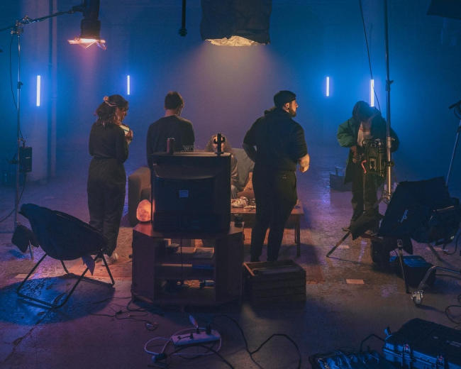 A group of five individuals in dark clothing is arranged around a low table in a dimly lit room with blue and purple lighting, setting up equipment for a project.