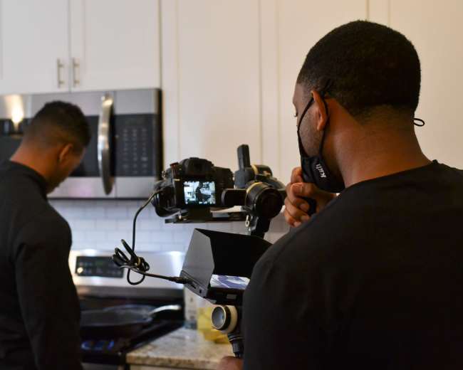 A person in a chef's jacket is cooking on a stovetop while another individual operates a camera and stabilizer in a kitchen setting.