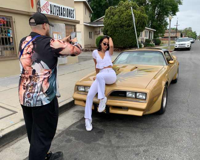 A photographer takes pictures of a model posing on a gold car on a street in front of a storefront.