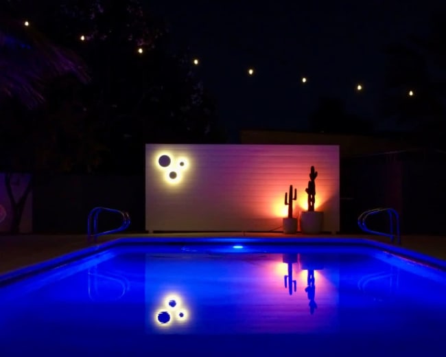 A swimming pool reflects colorful lights and a decorative wall with a cactus statue at night.